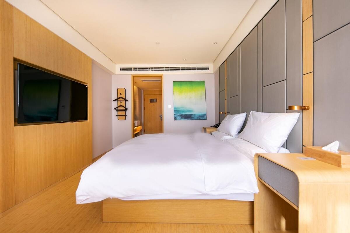 A modern hotel room with a large bed, a flat-screen TV mounted on the wall, a bedside table, and a painting on the wall.
