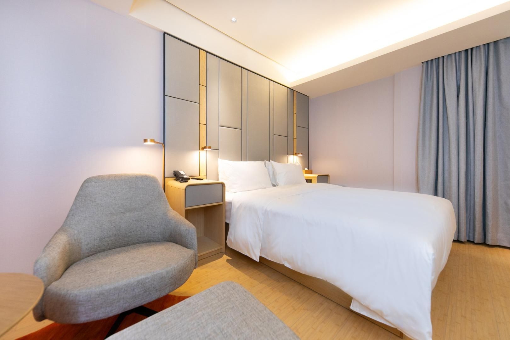 Une chambre d’hôtel moderne avec un grand lit, un fauteuil gris, des tables de chevet et une armoire intégrée.