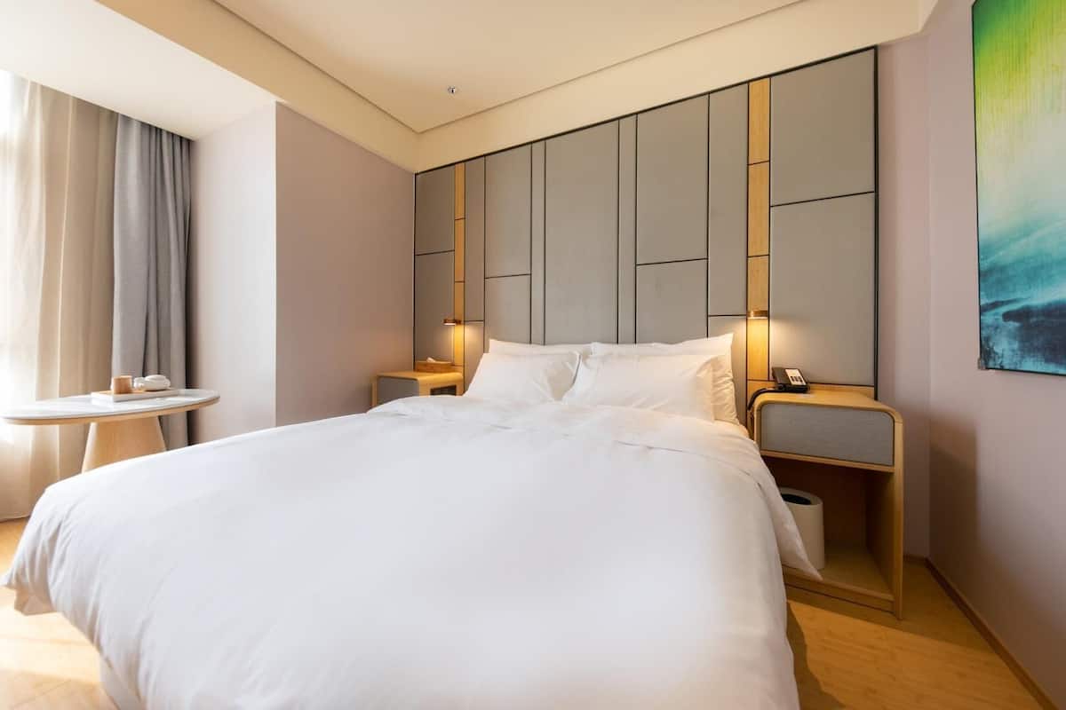 A modern hotel room with a large bed, bedside tables, a small round table, and a painting on the wall.
