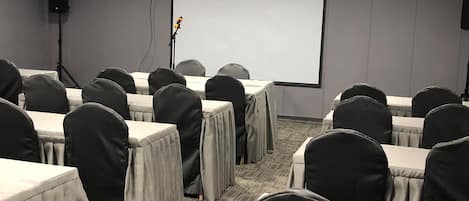 Uma sala de conferências com mesas, cadeiras, tela de projetor e um orador.