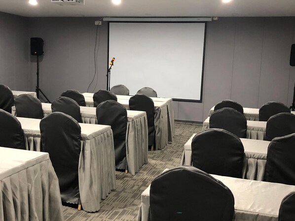 Un salón de conferencias con mesas, sillas, una pantalla para proyector y un orador.