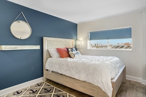 A bedroom with a bed, a round mirror, a window with blinds, and a view of buildings outside.