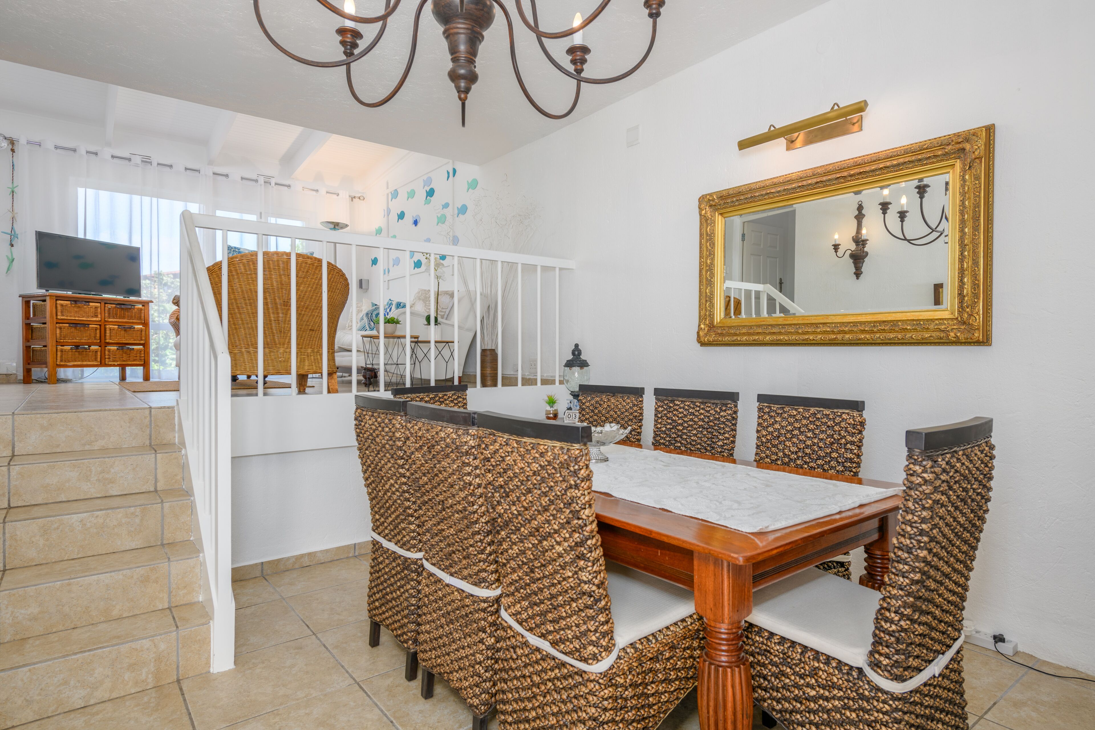 A dining area with a wooden table and wicker chairs, a chandelier, and a large mirror on the wall.