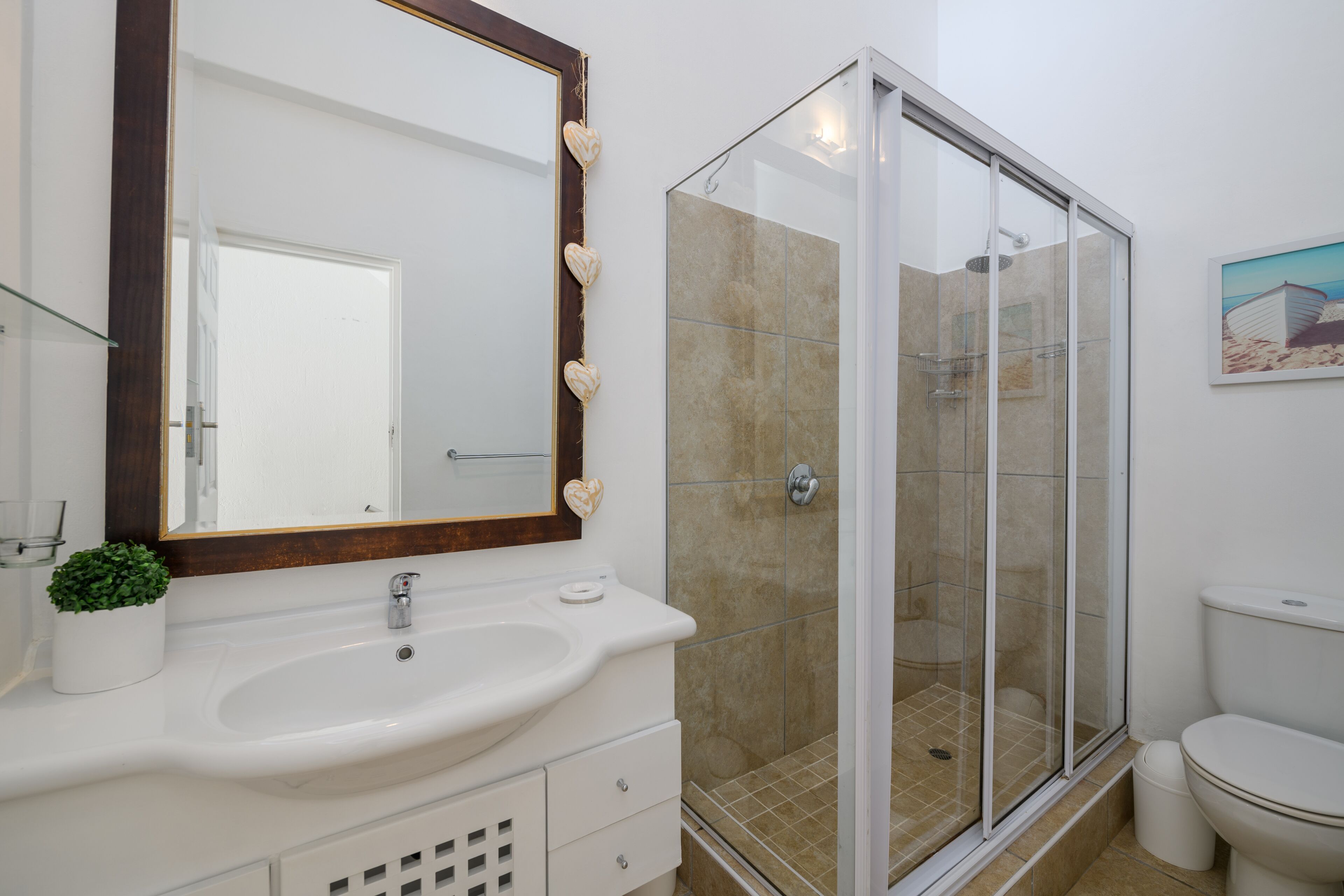 Een badkamer met een witte wasbak, een spiegel met houten lijst, een douchecabine van glas en een toilet.