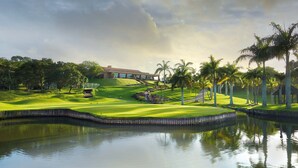 A golf course with a pond, palm trees, and a building in the background. - San Lameer Villa Rentals 3115 (Southbroom)