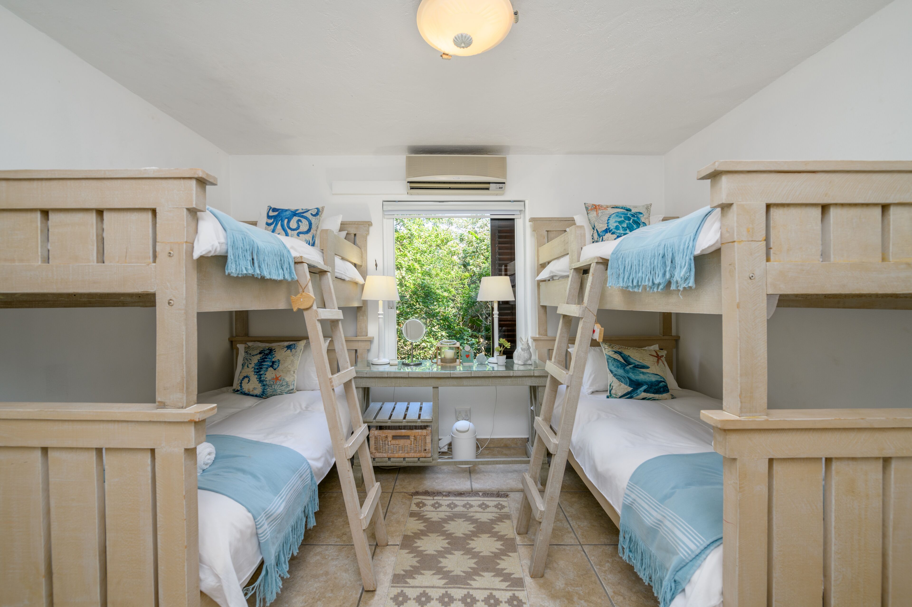 A room with bunk beds, a desk with a lamp, and a window with a view of greenery.