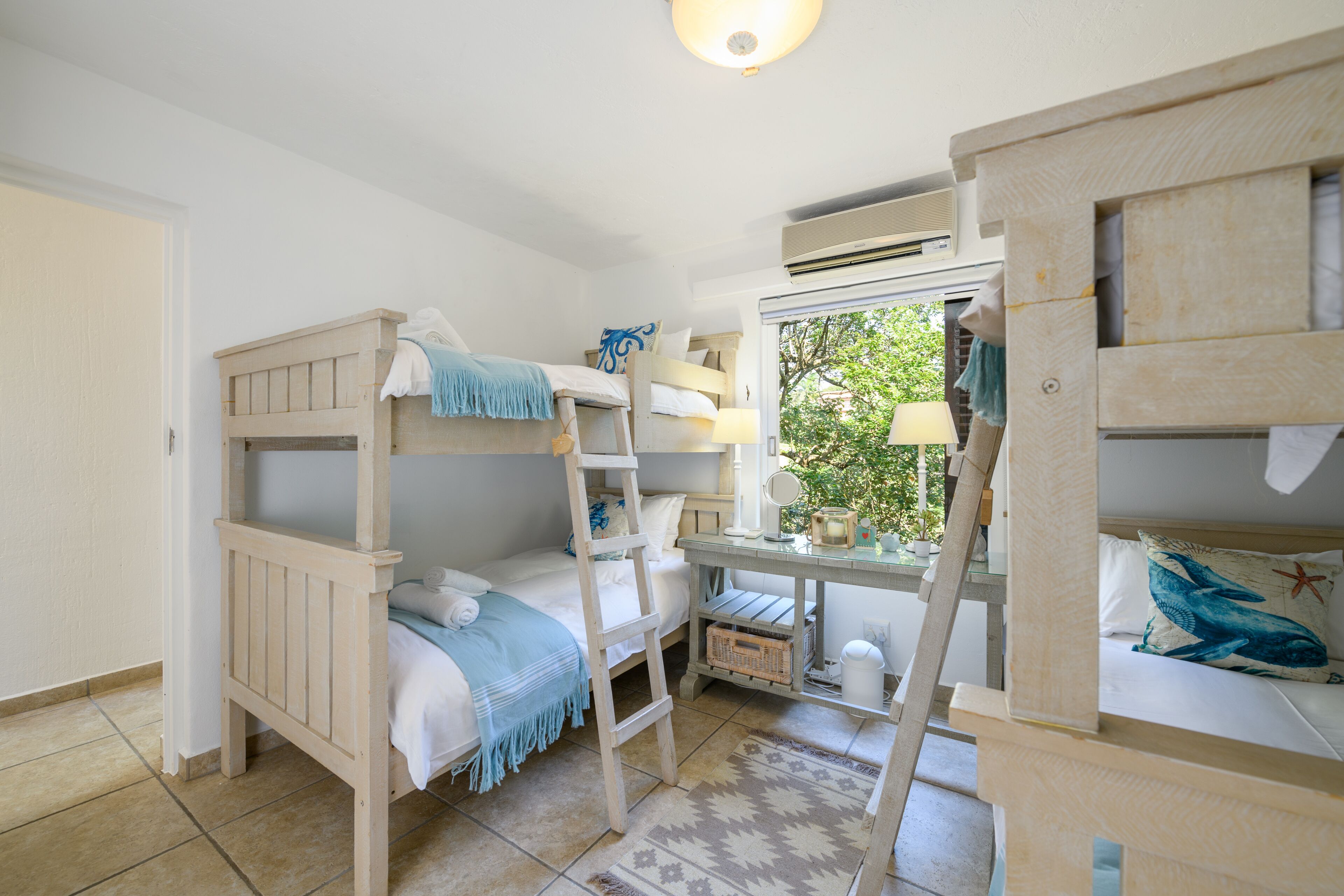 Habitación con literas, un escritorio, una escalera, una ventana con vistas a zonas verdes y una pequeña mesa con una lámpara.