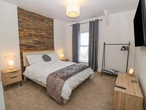 Quarto com cabeceira de madeira, cama com roupa de cama branca, duas luminárias de cabeceira, janela com cortinas, cômoda de madeira e televisão montada na parede.