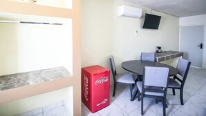 A dining area with a round table, four chairs, a red Coca-Cola cooler, a flat-screen TV mounted on the wall, and a white door. - OYO Hotel Plaza Las Torres, Cabo San Lucas (Cabo San Lucas)