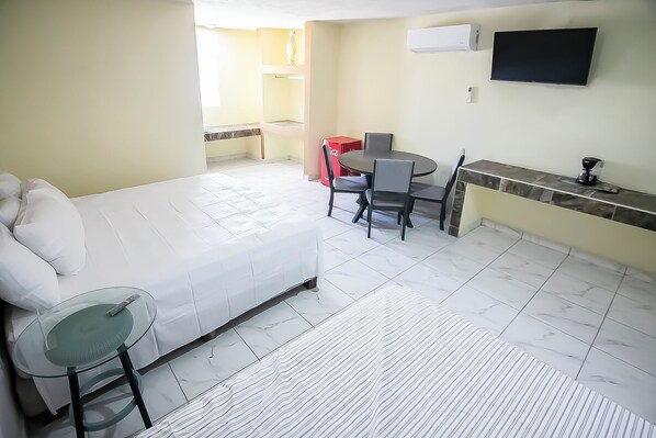 A hotel room with a bed, a glass table, a dining table with chairs, a red Coca-Cola cooler, a TV, and a tiled floor.