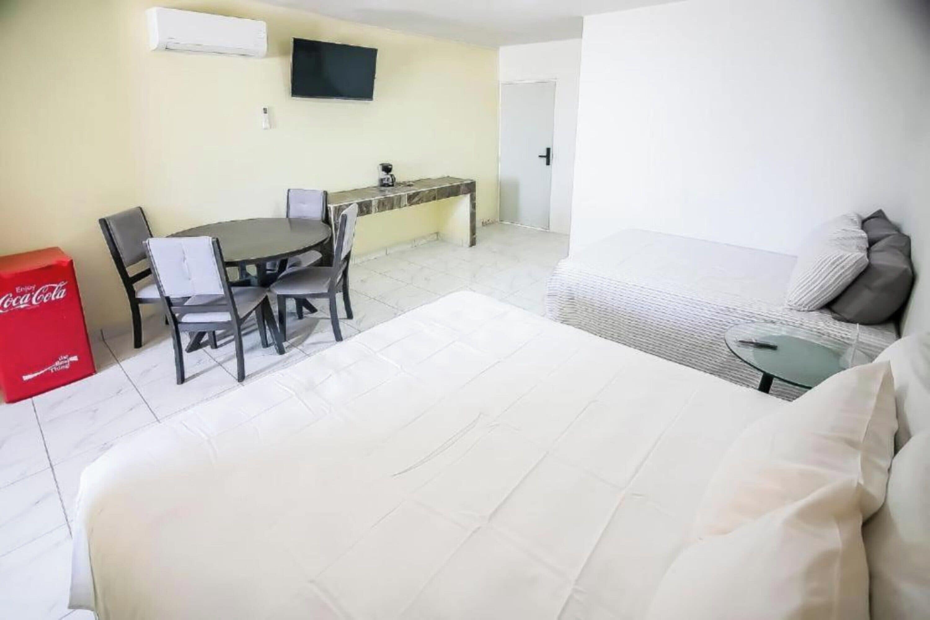 Una habitación de hotel con una cama, una mesa de comedor con sillas, una mesita, un sofá, un televisor montado en la pared y un refrigerador rojo Coca-Cola.