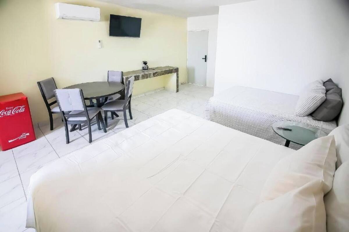 Ein Hotelzimmer mit Bett, Esstisch mit Stühlen, einem kleinen Tisch, einer Couch, einem an der Wand befestigten Fernseher und einem roten Coca-Cola-Kühler.