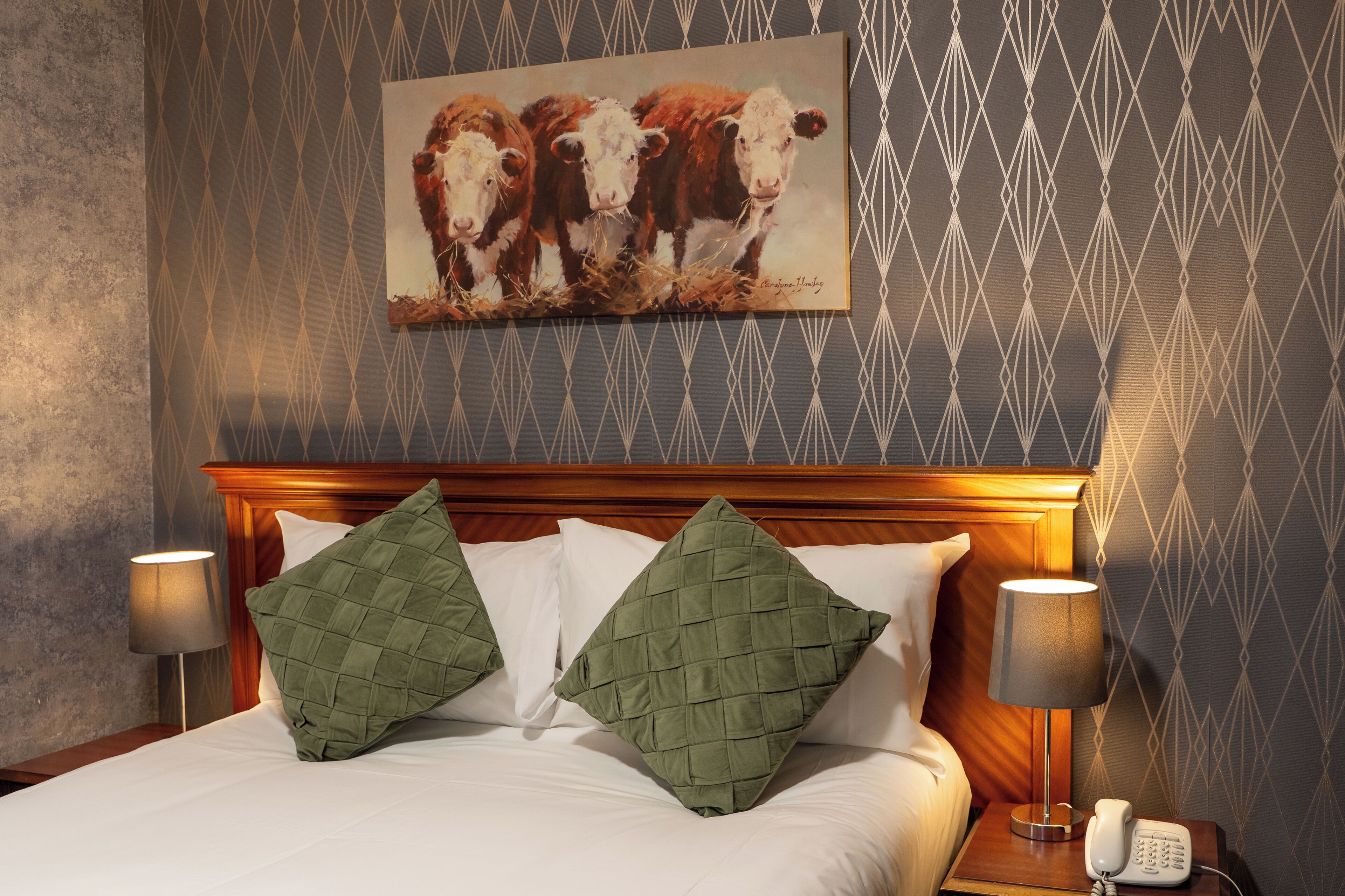 Une chambre d’hôtel avec un lit, deux oreillers verts, un tableau représentant des vaches accroché au mur et un téléphone sur la table de chevet.