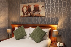 Habitación de hotel con una cama, dos almohadas verdes, un cuadro de vacas en la pared y un teléfono en la mesita de noche.