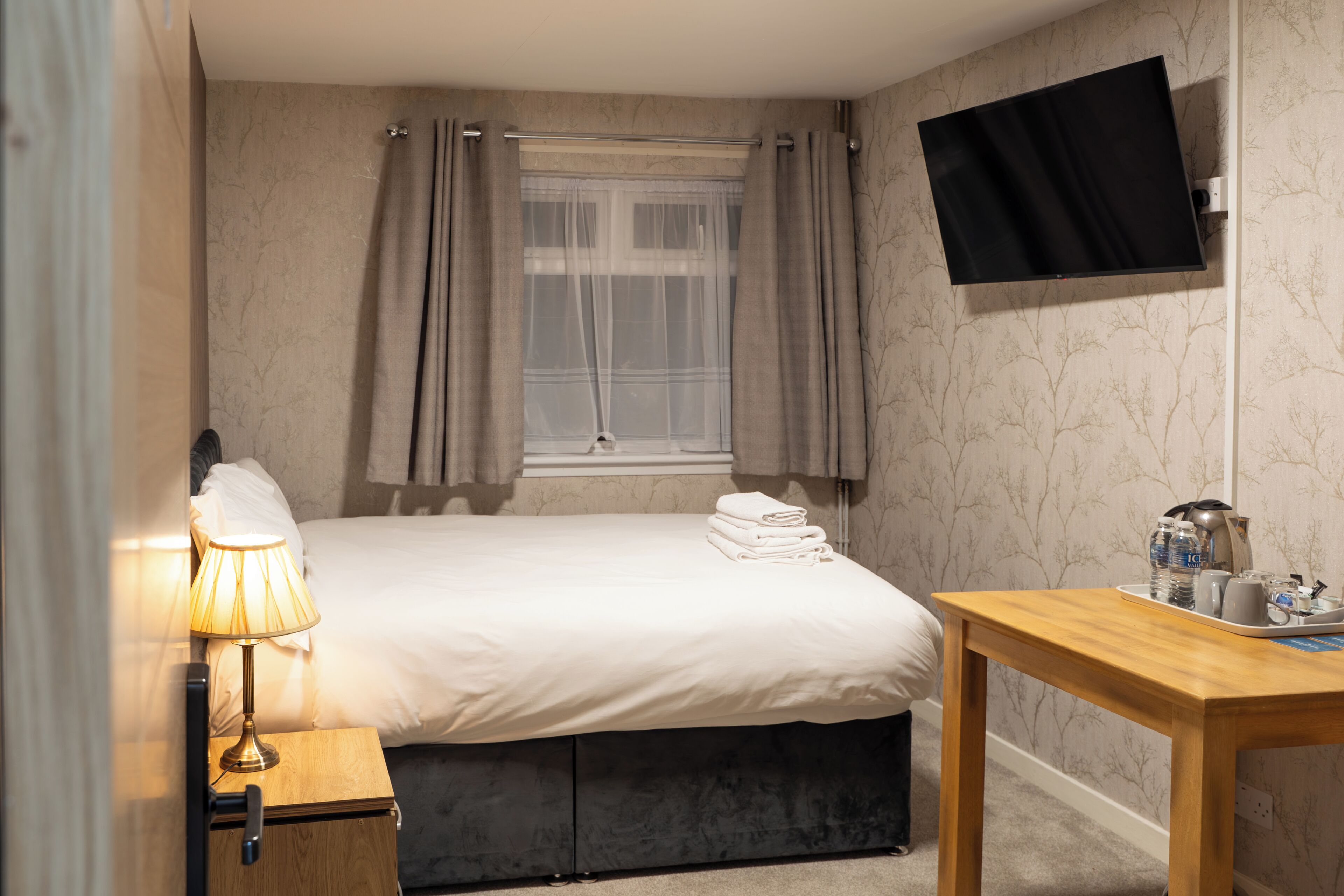 A hotel room with a bed, a bedside table with a lamp, a TV mounted on the wall, and a window with curtains.