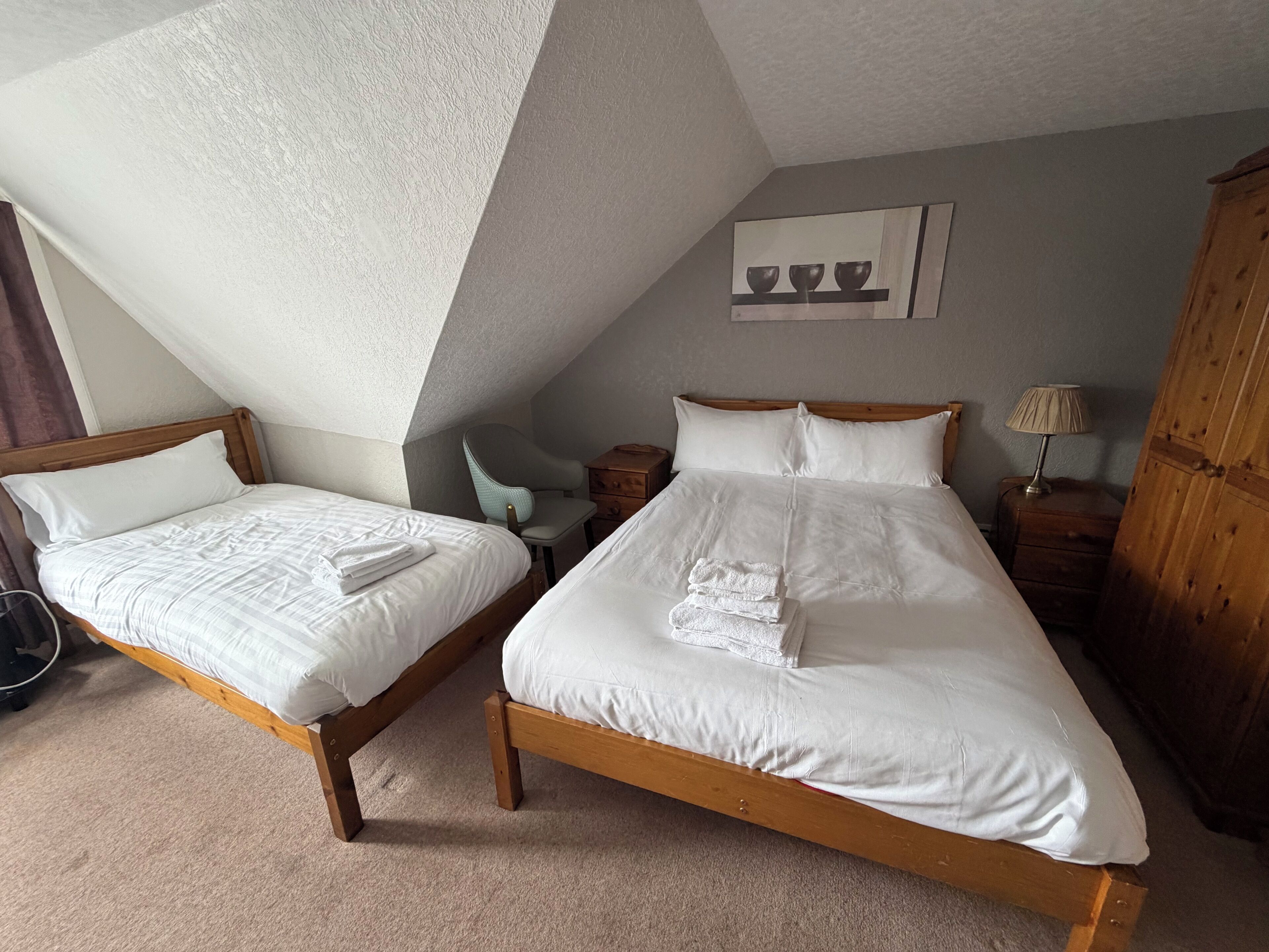 A room with two wooden beds, a chair, a nightstand, a lamp, and a cabinet.