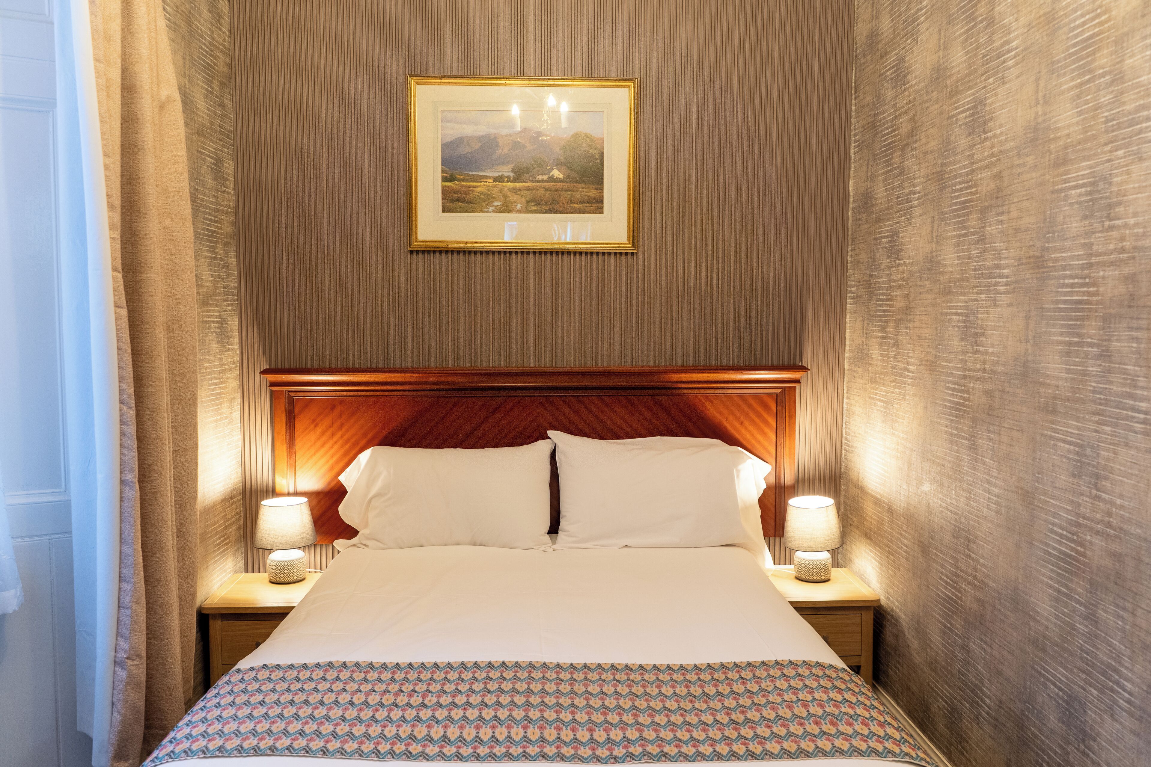 A hotel room with a large bed, two bedside lamps, a wooden headboard, and a framed picture on the wall.