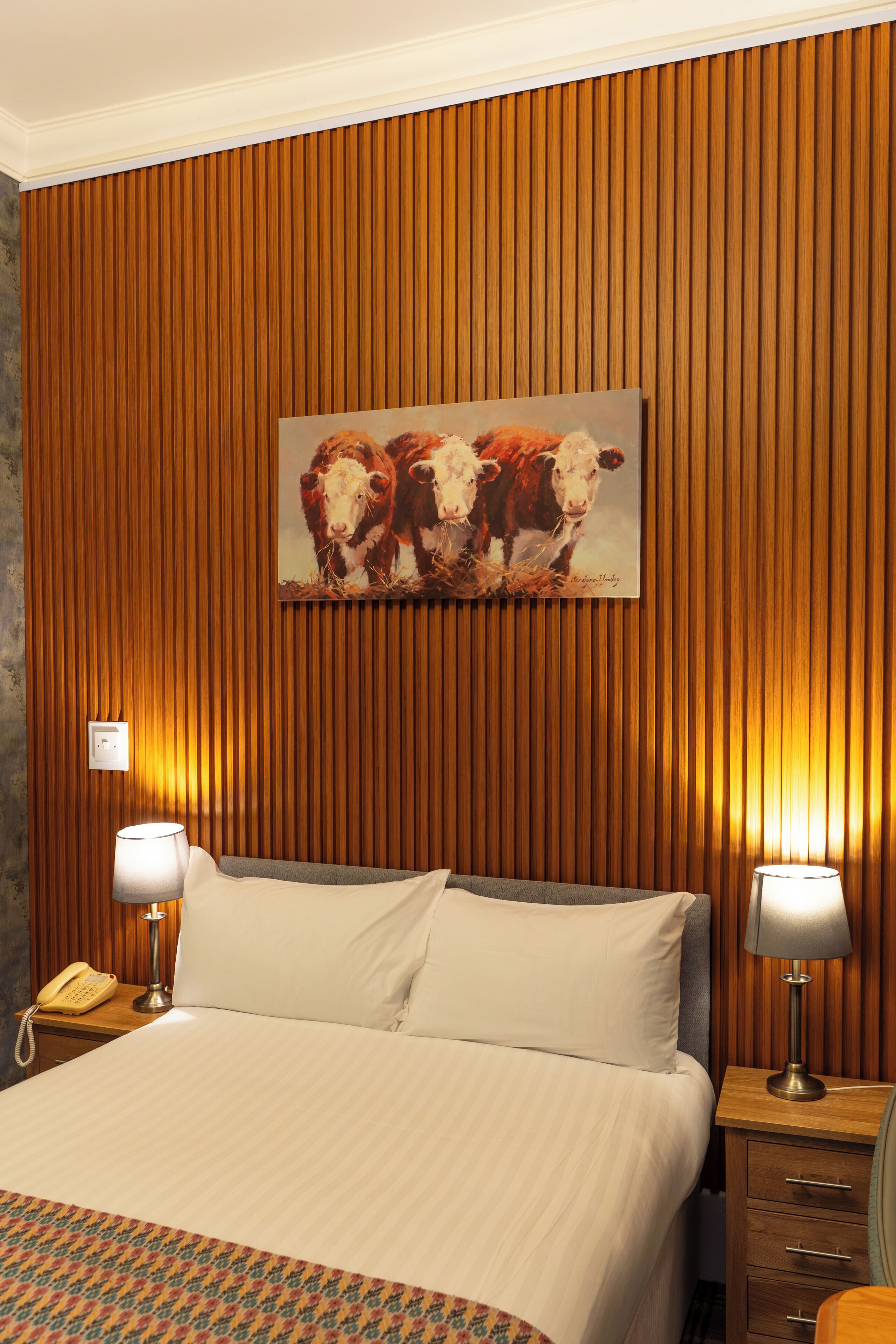 Een hotelkamer met een bed, twee bedlampen, een schilderij van koeien en houten lambrisering.