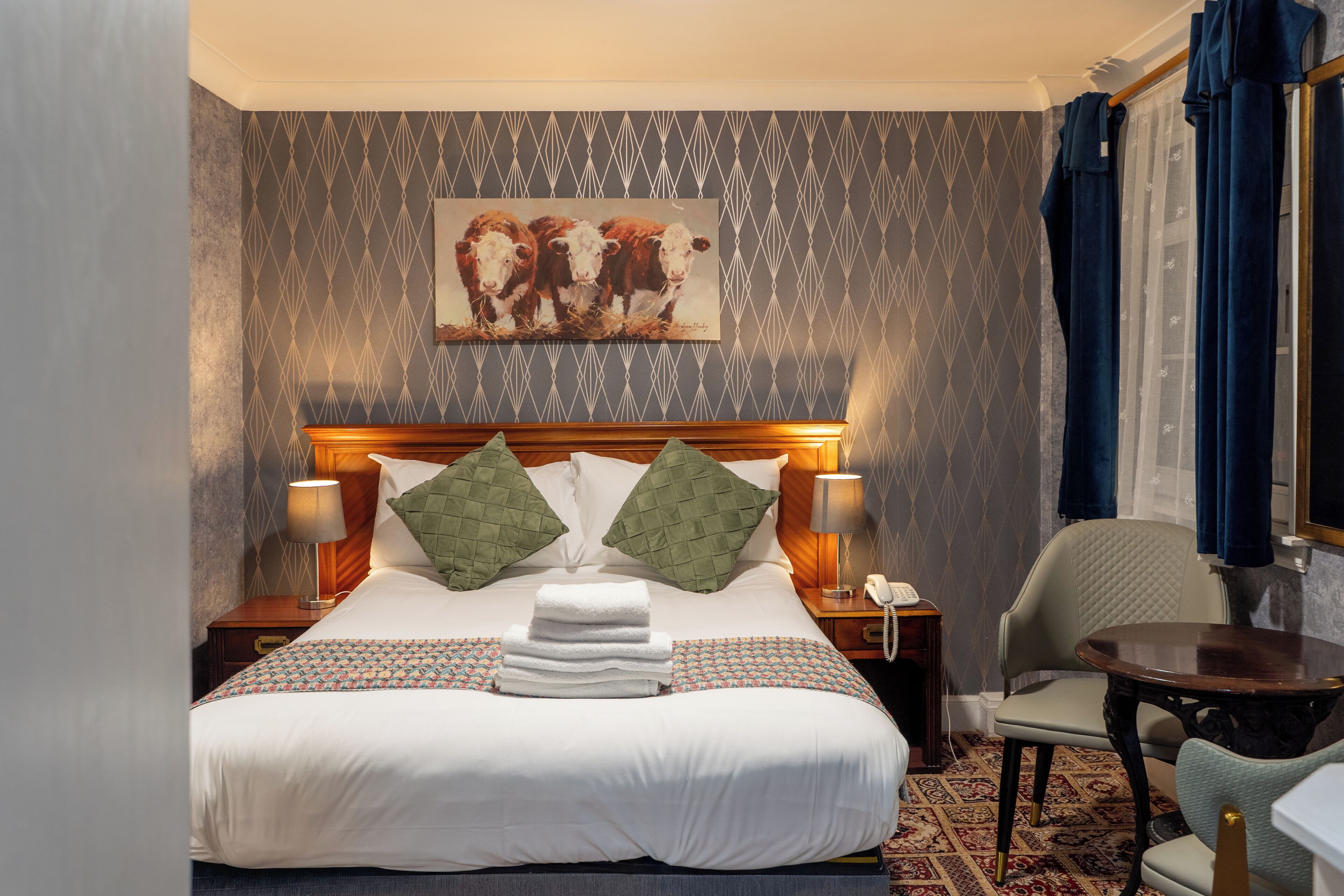 A hotel room with a bed, two pillows, a bedside lamp, a telephone, a chair, a small table, and a painting on the wall.
