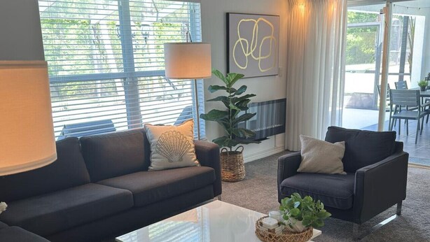 Una sala de estar moderna con un sofá, un sillón y una mesa de centro. Hay una chimenea, un ventanal grande y una planta sobre la mesa de centro.