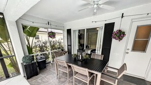 Un porche cubierto con una mesa, sillas, ventilador de techo y cestas de flores colgantes.