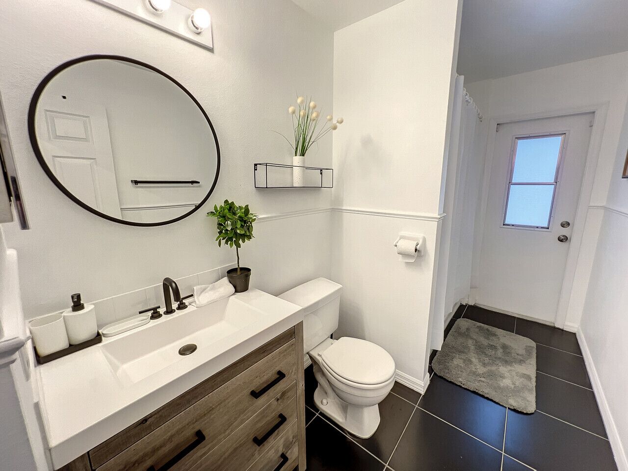 Ein modernes Badezimmer mit einem weißen Waschbecken, einem Spiegel und einer Toilette. Auf dem Waschtisch steht eine kleine Pflanze und auf dem Boden liegt ein Teppich.