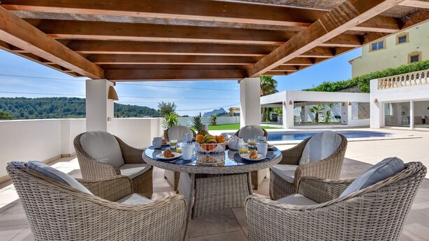 Un espace extérieur abrité avec des meubles en osier, une table où sont disposés des aliments pour le petit-déjeuner, et une piscine en arrière-plan.