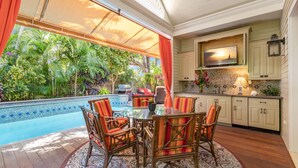 Un área de comedor junto a la piscina con sillas de mimbre y una mesa de vidrio, rodeada por una piscina y abundante vegetación.