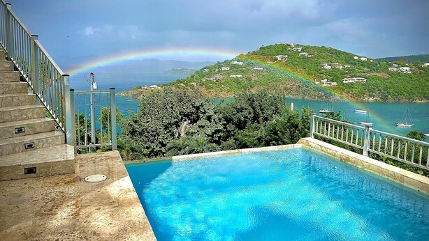 Una piscina con vista sul paesaggio costiero, inclusi un arcobaleno e case su una collina.