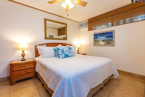 A hotel room with a large bed, two bedside tables with lamps, a ceiling fan, a wall-mounted air conditioner, a large mirror, and a framed picture on the wall.