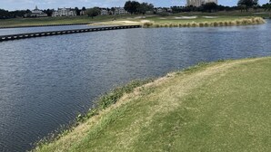 A calm lake with a grassy shoreline, a distant building complex, and a clear blue sky with clouds. - Golfer’s heaven, Orlando with 3 Champ courses (Kissimmee)