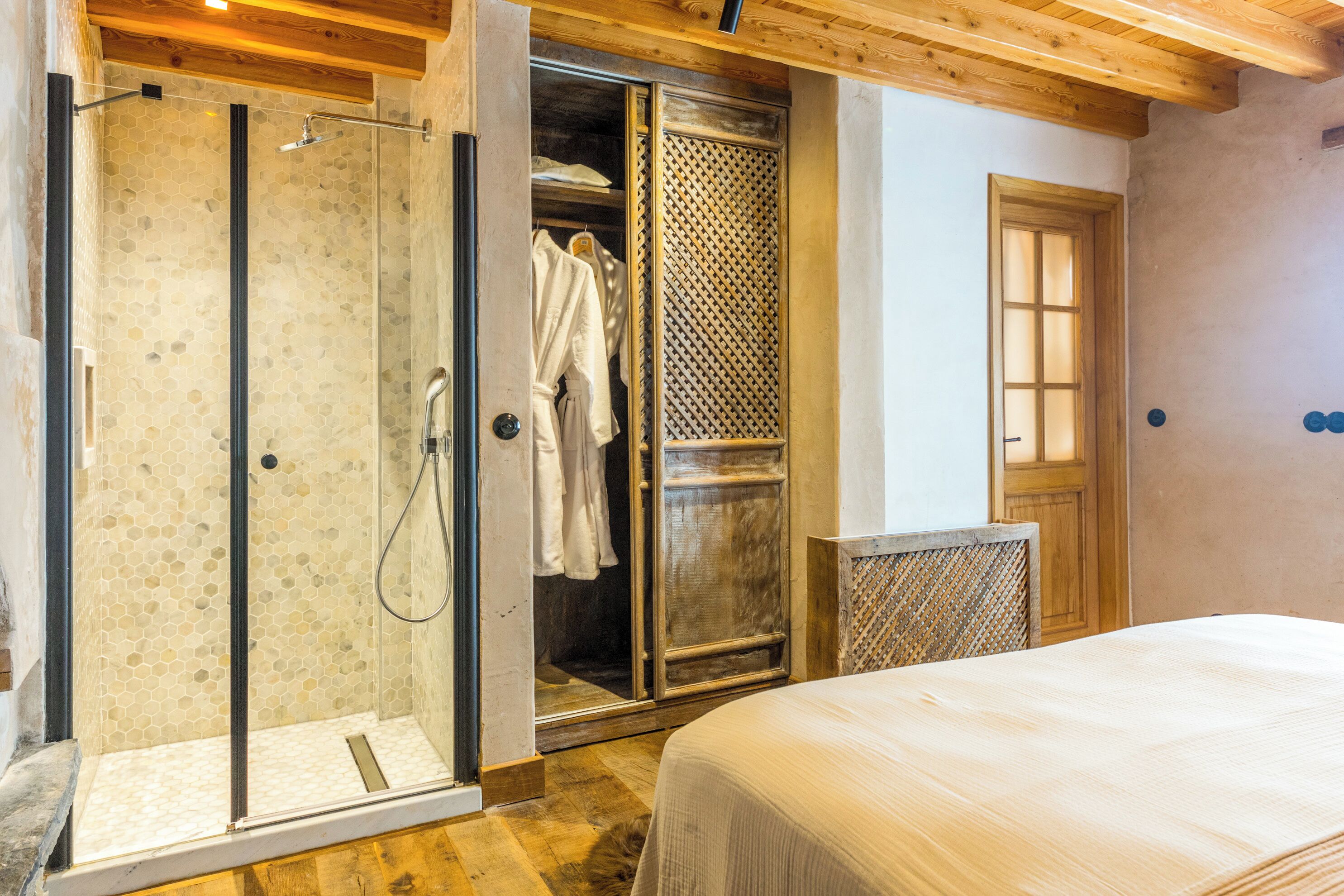 A bedroom with a large bed, a walk-in closet with hanging clothes, and a shower area with a glass door.