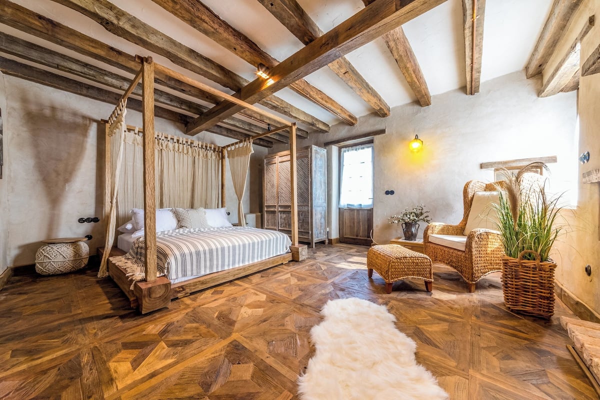 Une chambre à coucher rustique avec un lit à baldaquin, des poutres apparentes et un parquet disposé en motif chevrons.