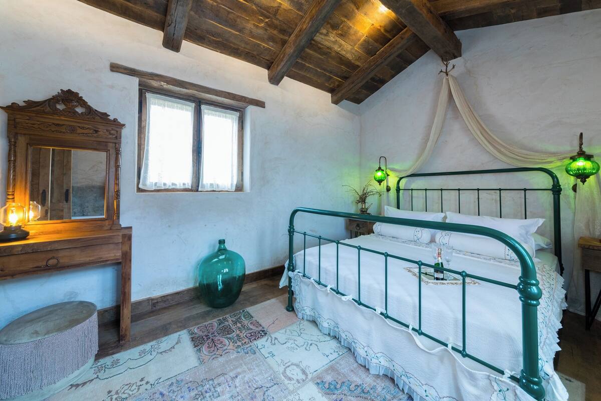 Une pièce avec un plafond en bois, un cadre de lit en métal vert, un grand miroir et une fenêtre avec des rideaux.