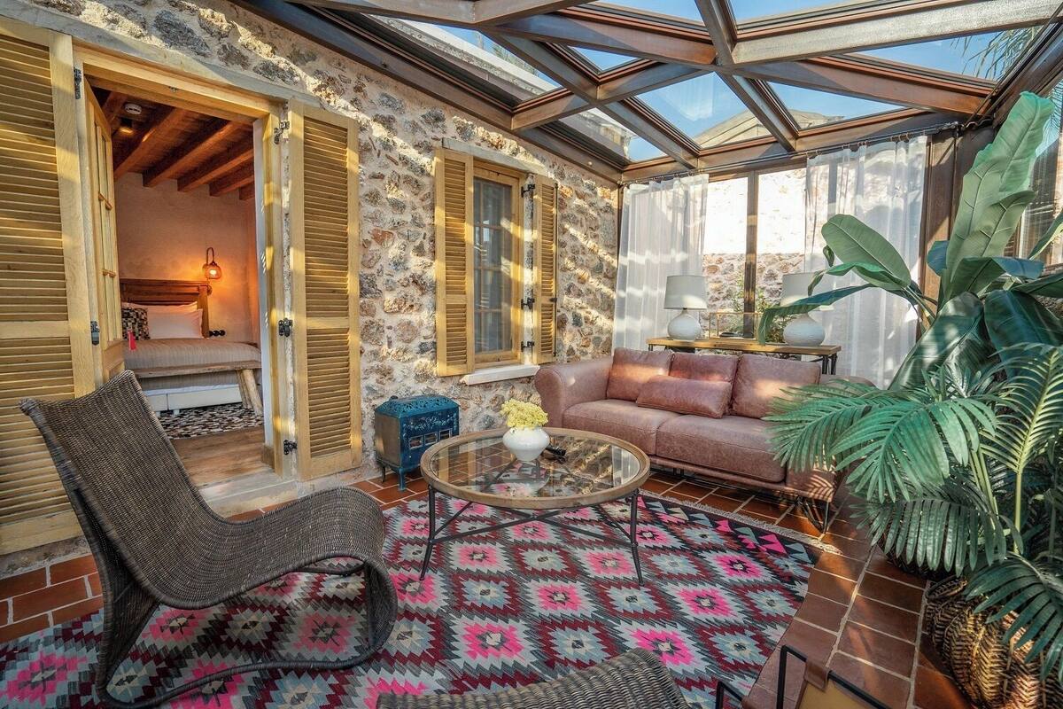 Une pièce chaleureuse avec un mur en pierre, des volets en bois, un plafond en verre, un canapé, un fauteuil, une petite table et un tapis à motifs.