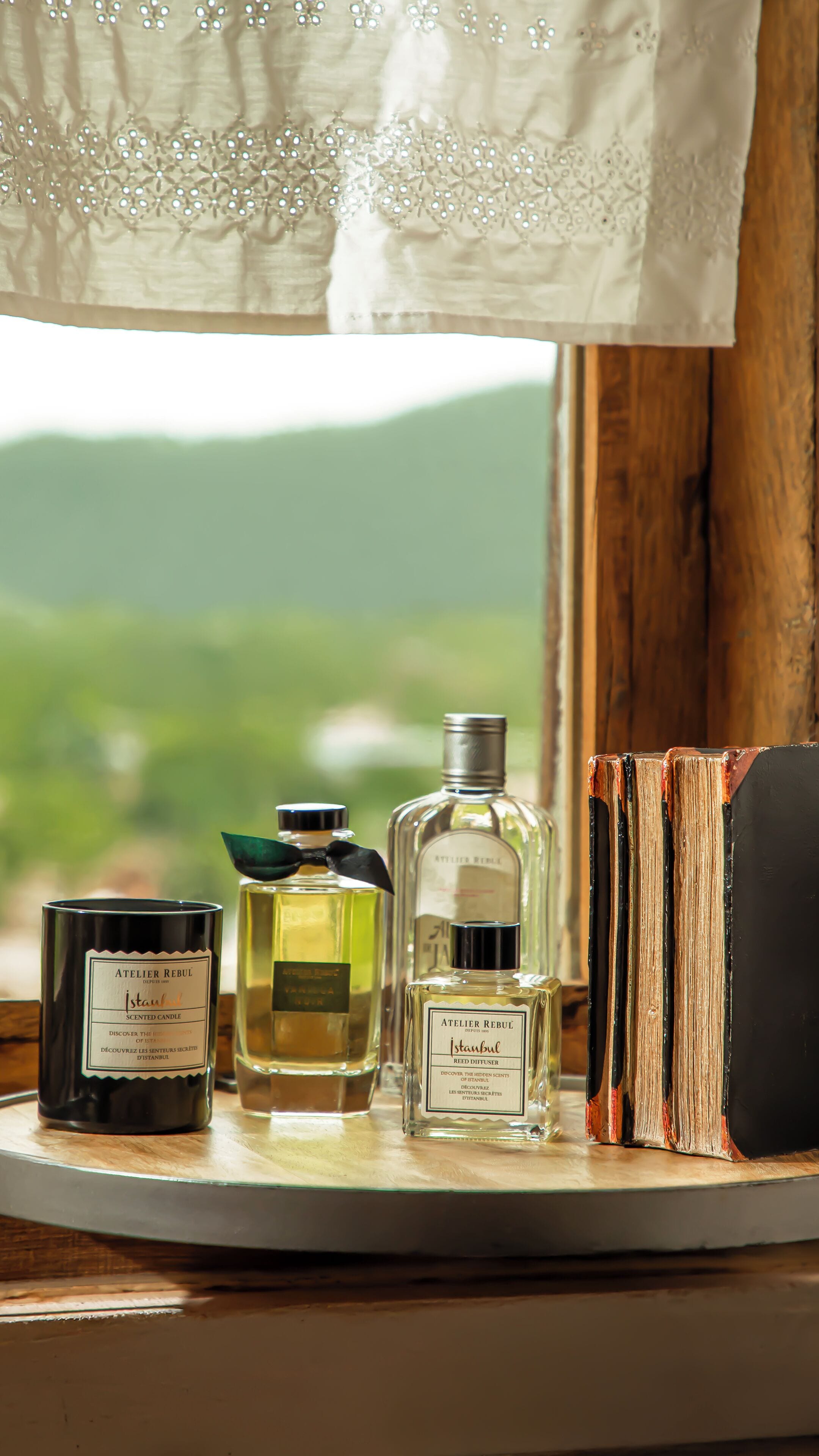 A table with three perfume bottles and a candle, a book, and a window with a view of greenery.
