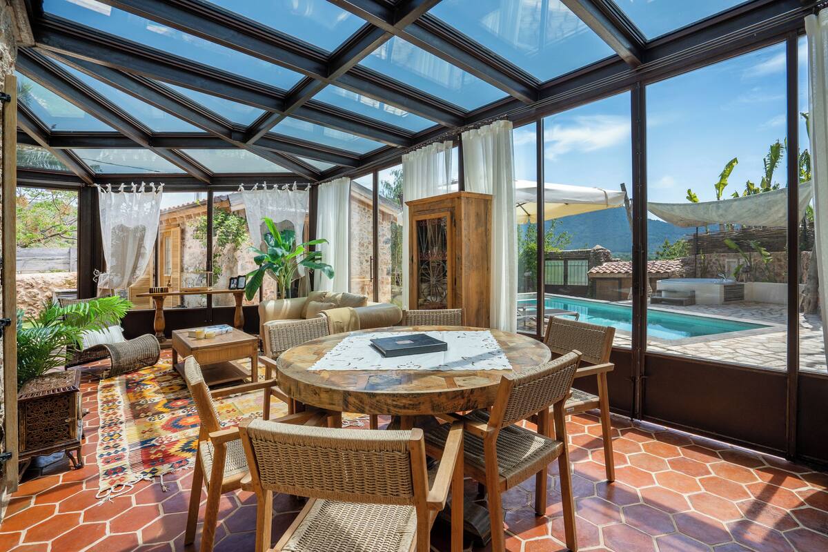 Une terrasse ensoleillée avec une table ronde en bois, des chaises en osier et une vue sur une piscine et des montagnes.