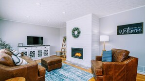 A living room with leather sofas, a fireplace, a television, and a welcome sign. - Homestead @ Oakley Farms (Cumming)