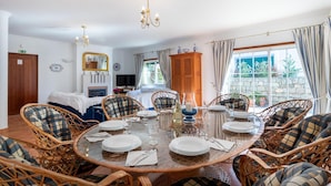 A dining area with a round table set for six, surrounded by wicker chairs with plaid cushions. A chandelier hangs from the ceiling, and a wooden cabinet is visible in the background. - Villa Goshen - Praia da Luz (Luz)