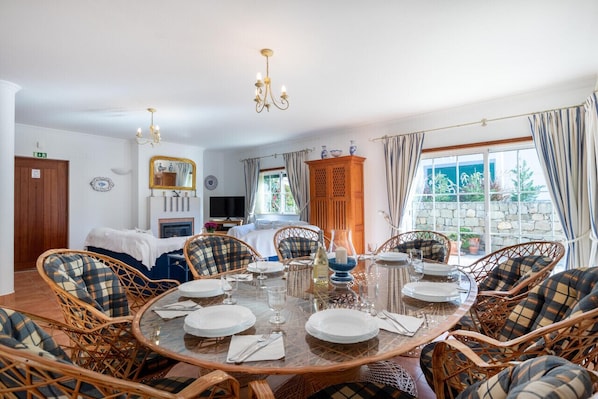 A dining area with a round table set for six, surrounded by wicker chairs with plaid cushions. A chandelier hangs from the ceiling, and a wooden cabinet is visible in the background. - Villa Goshen - Praia da Luz (Luz)