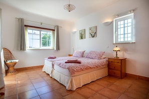 A bedroom with two beds, a wicker chair, a bedside table with a lamp, and a window with curtains. - Villa Goshen - Praia da Luz (Luz)
