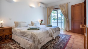 A bedroom with two beds, a nightstand, a lamp, a window with curtains, and a door. - Villa Goshen - Praia da Luz (Luz)