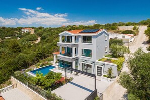 A modern two-story house with a swimming pool, surrounded by greenery and a clear sky.
