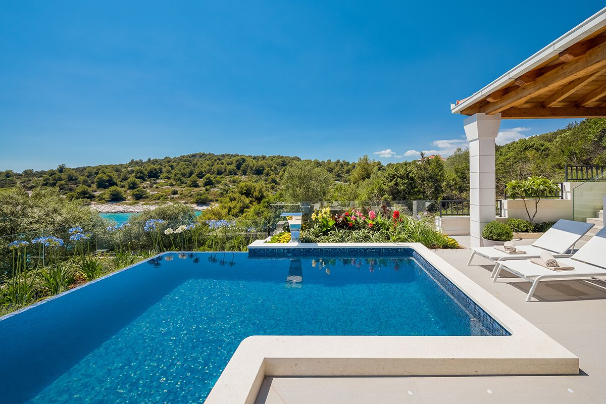 Ein rechteckiger Swimmingpool mit klarem, blauem Wasser, umgeben von einer weißen Liegefläche. Weiße Liegestühle und ein kleiner Gartenbereich mit bunten Blumen sind ebenfalls vorhanden.