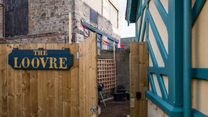 Una puerta de madera con un letrero que dice “The Loovre” da acceso a un camino de ladrillo y un edificio con paredes de piedra vista y detalles azules.