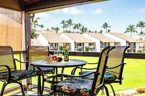 A covered outdoor seating area with a table and chairs, overlooking a lawn with palm trees and hotel buildings in the background.