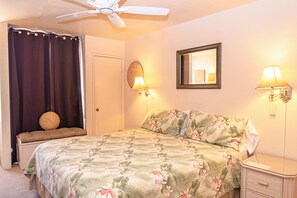 A bedroom with a bed, a ceiling fan, a bedside table, a nightstand, a lamp, a mirror, and a window with curtains.