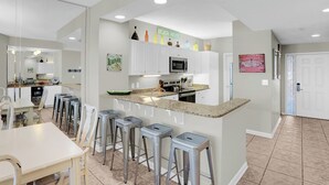 A modern kitchen with white cabinets, a granite countertop, and a microwave. There are bar stools and a dining table.
