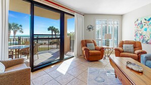A spacious living room with a sliding glass door leading to a balcony, furnished with leather chairs and a wooden coffee table.