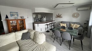A modern living room with a glass dining table, grey sofas, and a kitchen in the background.